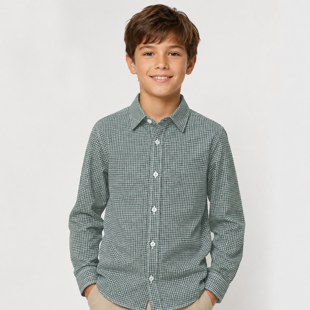 A young boy smiling and wearing a long-sleeve button-up shirt with a fine green and white micro-check pattern, paired with beige pants, against a light gray studio background.