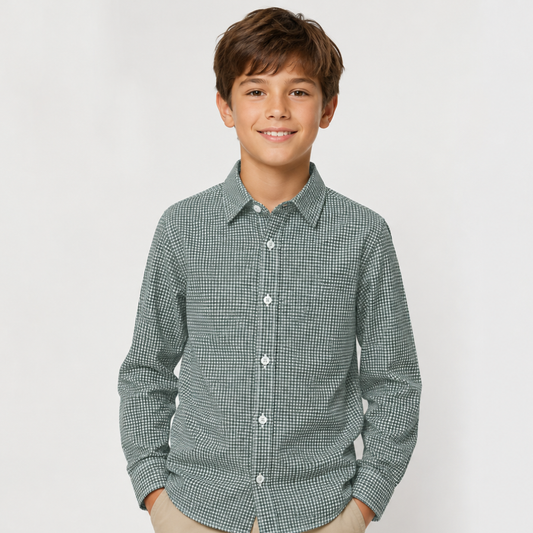 A young boy smiling and wearing a long-sleeve button-up shirt with a fine green and white micro-check pattern, paired with beige pants, against a light gray studio background.