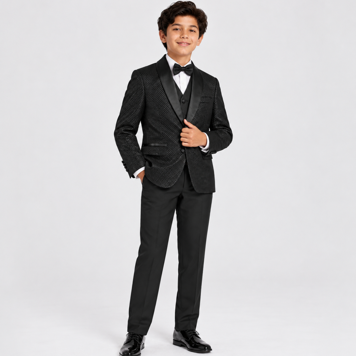 A young boy modeling a black formal kids' tuxedo suit with a textured pattern on the jacket, black satin lapels, a matching vest, black trousers, a white dress shirt, and a black bow tie, standing against a light gray studio background.