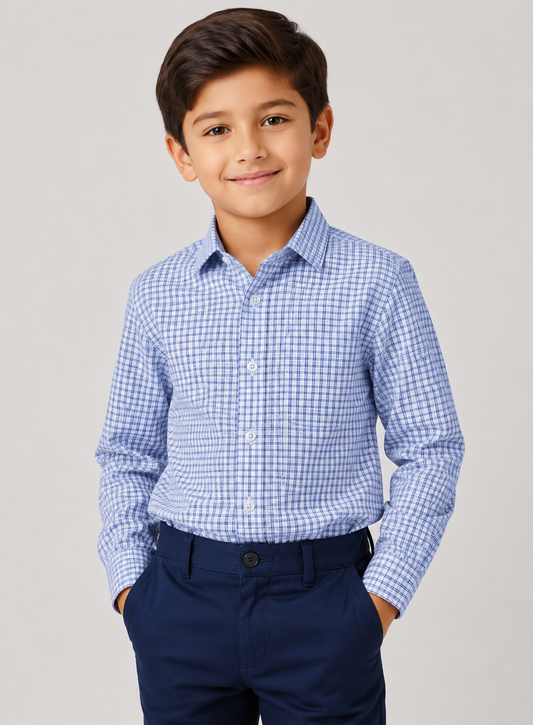 A young boy wearing a blue and white checkered long-sleeve button-down shirt with dark navy trousers, standing against a light gray studio background.