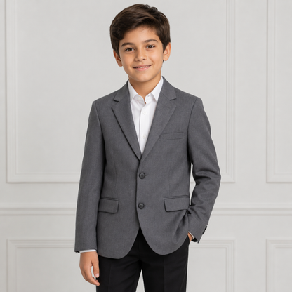A young boy wearing a gray two-button blazer over a white collared shirt and black trousers, standing against a white paneled wall.