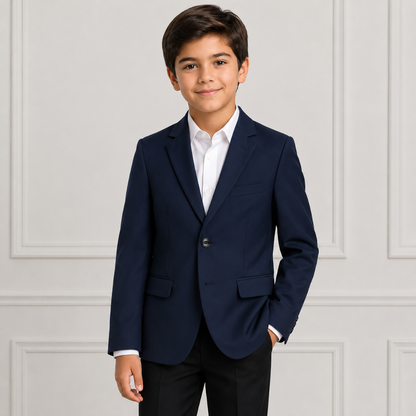 A young boy wearing a navy blue boys' blazer over a white dress shirt and black trousers, standing against a light gray paneled wall.