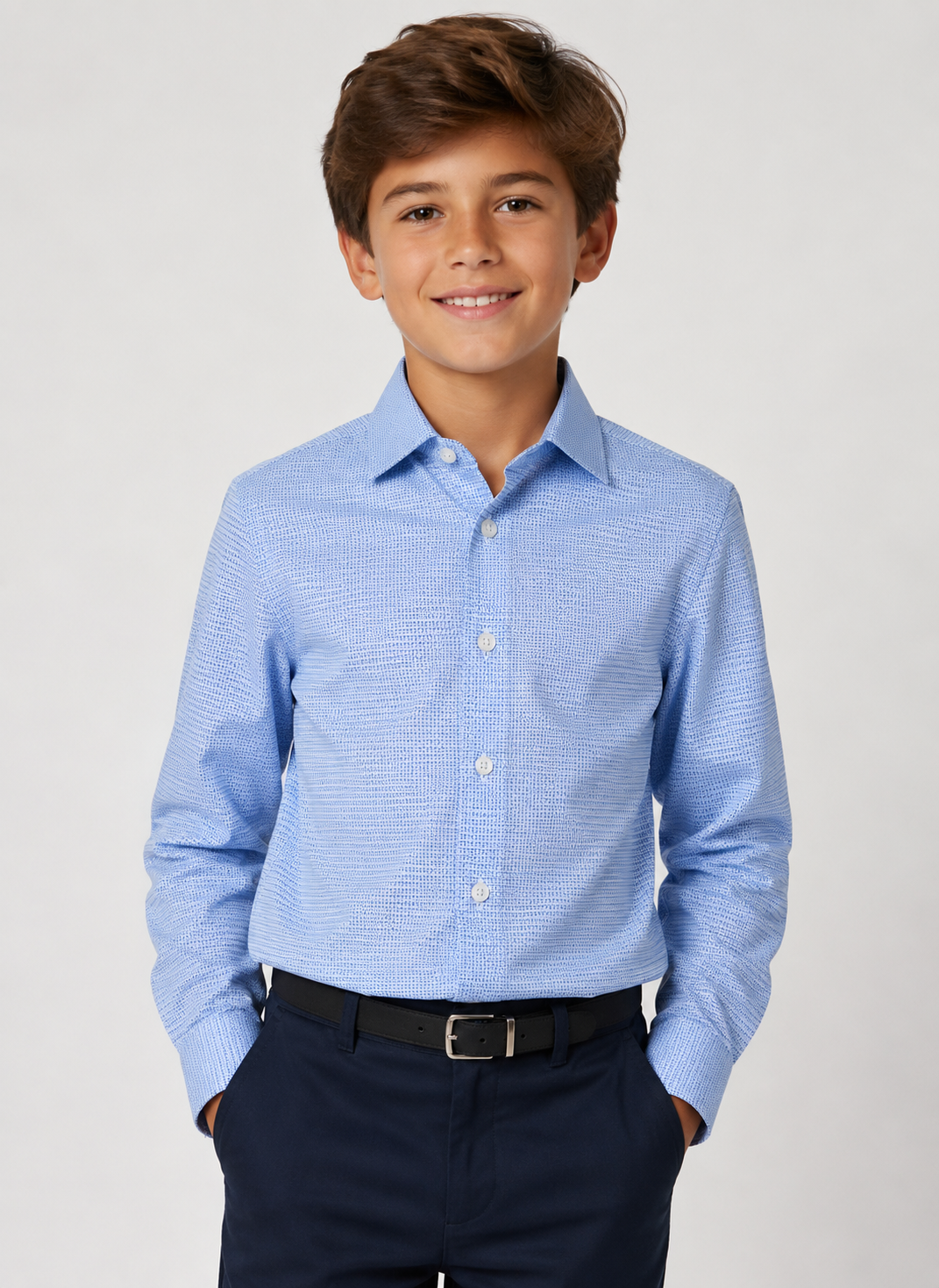 A young boy smiling, wearing a light blue long-sleeve button-down shirt with a subtle textured pattern, paired with navy pants and a black belt, against a light gray studio background.