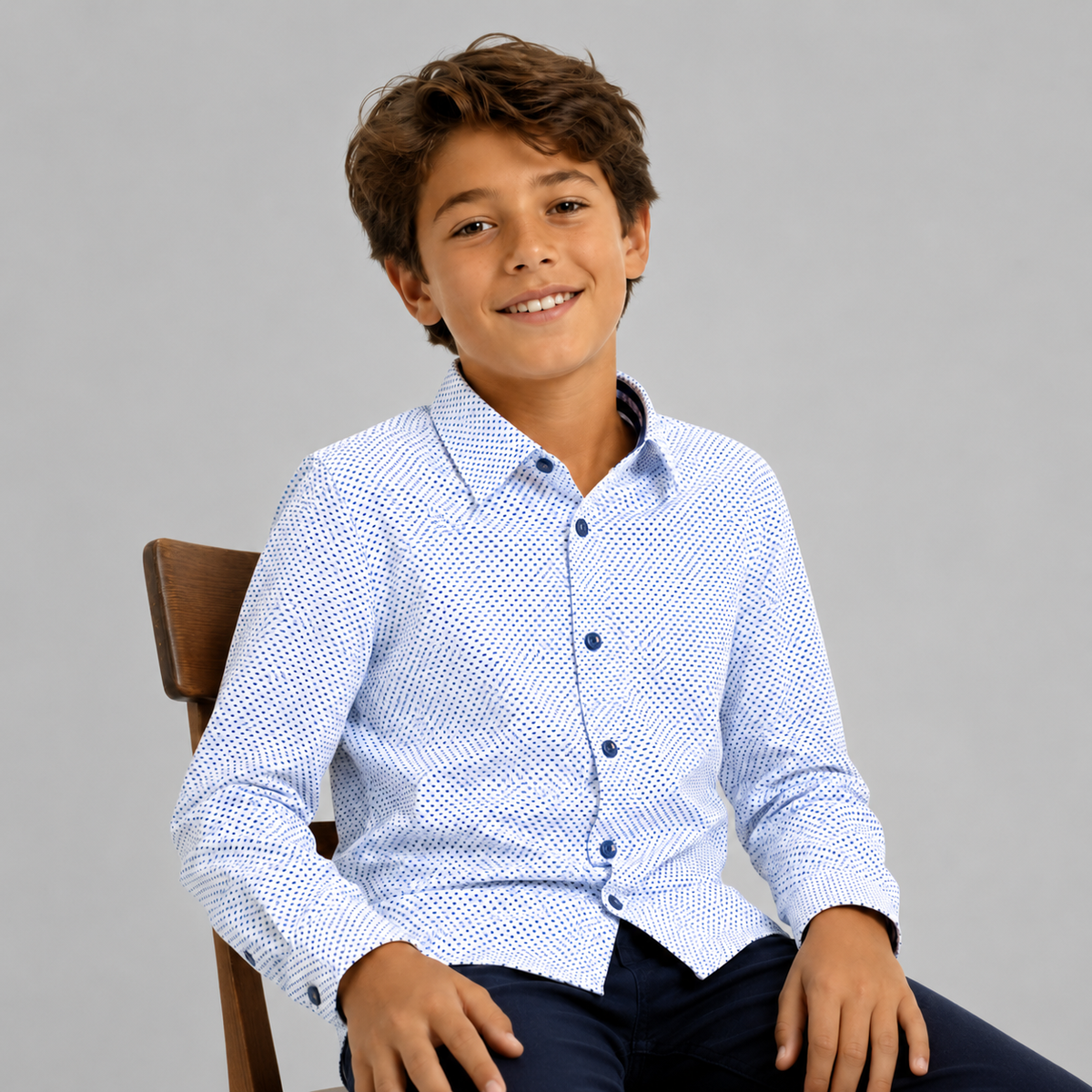 Young boy wearing a blue patterned shirt sitting on a chair against a gray background