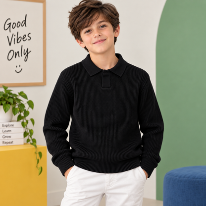 A young boy wearing a black textured waffle-knit long-sleeve polo sweater and white pants, standing in a brightly lit room with a green wall, a yellow side table, and a framed sign that reads 'Good Vibes Only'.
