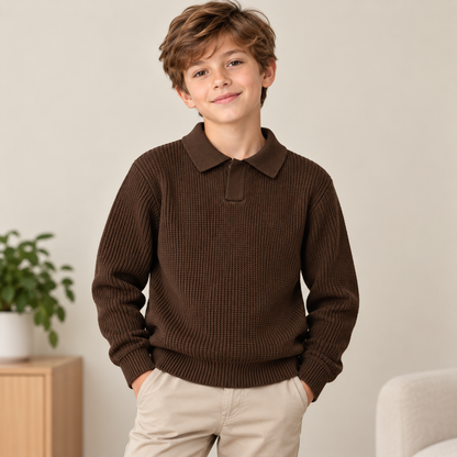 A young boy wearing a dark brown ribbed knit polo sweater and beige pants, standing in a brightly lit indoor room with a light beige wall, a wooden side table with a green plant, and a light-colored chair.