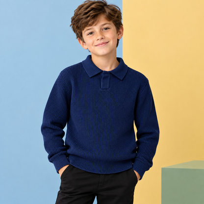 A young boy with brown hair smiling, wearing a navy blue ribbed knit polo sweater and black pants, standing against a split blue and yellow background.