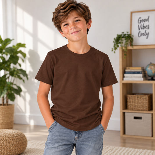 A young boy with wavy brown hair smiling in a bright room, wearing a solid dark brown short-sleeve crewneck t-shirt and light blue jeans.