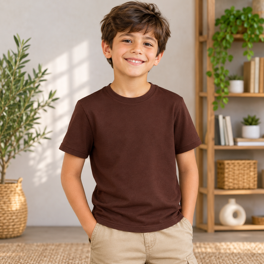 A smiling young boy wearing a solid dark brown short-sleeve crewneck t-shirt and khaki cargo shorts, standing in a brightly lit room with a wooden bookshelf and potted plants in the background.