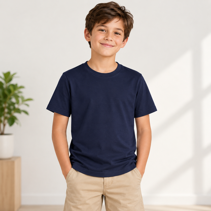 A young boy wearing a navy blue crewneck t-shirt and khaki pants, standing in a brightly lit room with a white wall and a small potted plant in the background.