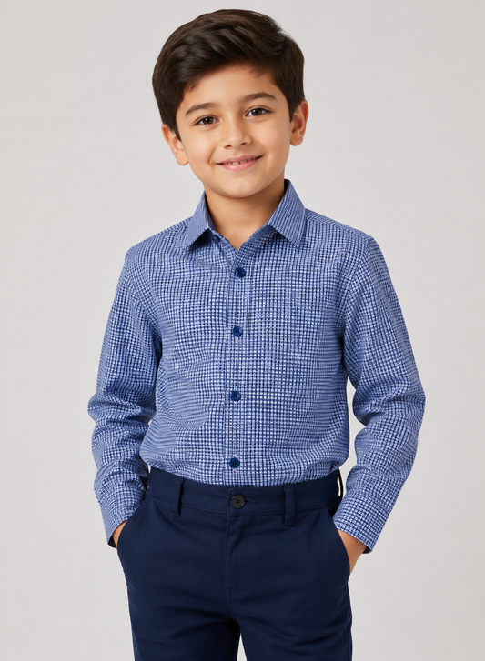 A young boy wearing a blue and white micro-check patterned long-sleeve button-down shirt with dark navy trousers, standing against a light gray studio background.