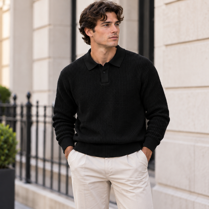 A man wearing a black textured knit polo sweater and off-white trousers, standing outdoors in front of a light-colored stone building.