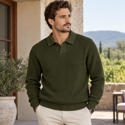A man wearing an olive green ribbed knit polo sweater and cream trousers, standing outdoors on a patio with a stone wall, greenery, and distant hills in the background.