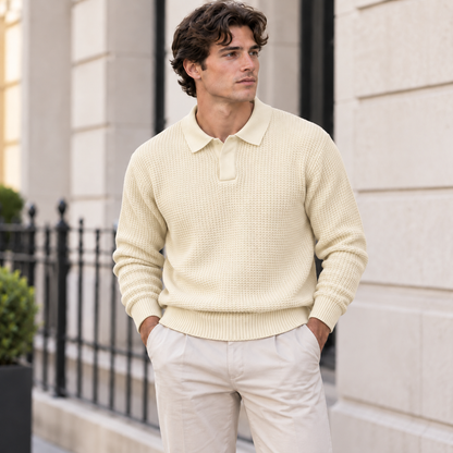 A man wearing a cream-colored waffle-knit polo sweater and light beige trousers, standing outdoors in front of a light-colored stone building.