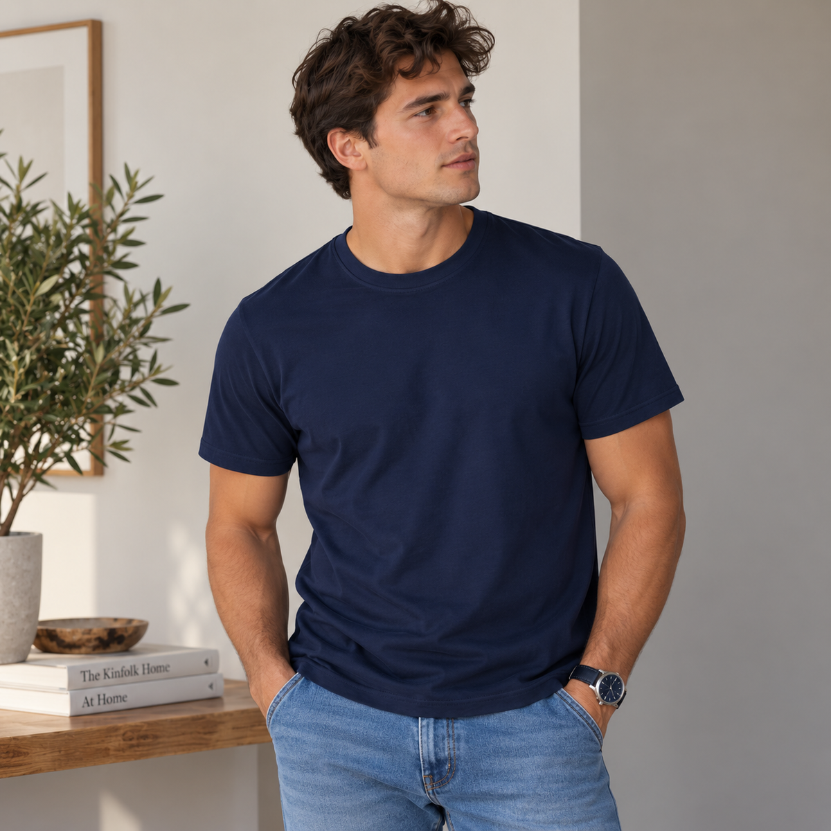 A man wearing a navy blue crewneck t-shirt and light blue jeans, standing in a modern indoor setting with a wooden console table, books, and a potted plant in the background.